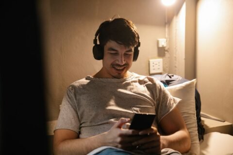 Smiling man in a gray T-shirt wearing black headphones and watching videos on his smartphone while relaxing indoors