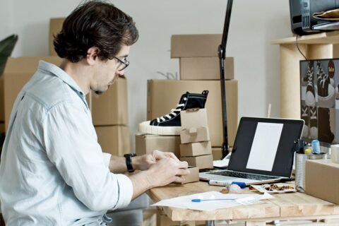 Man in glasses packing sneakers into cardboard boxes at a desk with a laptop for an online store
