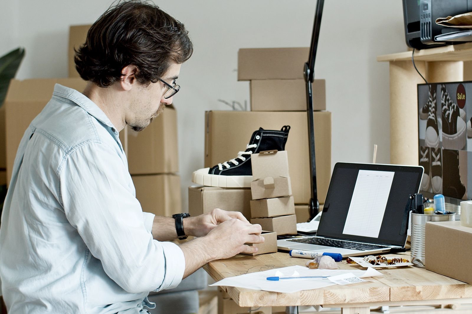 Man in glasses packing sneakers into cardboard boxes at a desk with a laptop for an online store