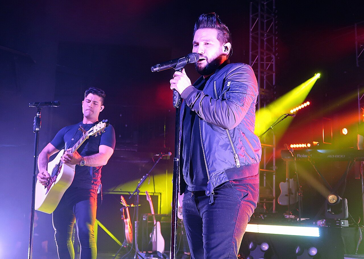 Country duo Dan + Shay on stage in 2017, one singing into a microphone and the other playing acoustic guitar under stage lights