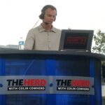 Broadcaster Colin Cowherd wearing a headset at an outdoor radio desk branded The Herd with Colin Cowherd, 2010