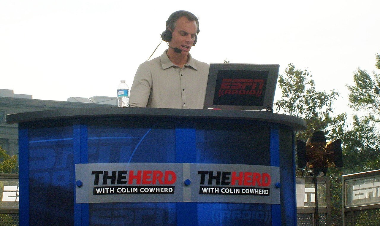 Broadcaster Colin Cowherd wearing a headset at an outdoor radio desk branded The Herd with Colin Cowherd, 2010