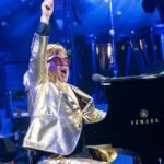 Elton John in a gold jacket raising one arm while performing at a piano on stage at Glastonbury in 2023