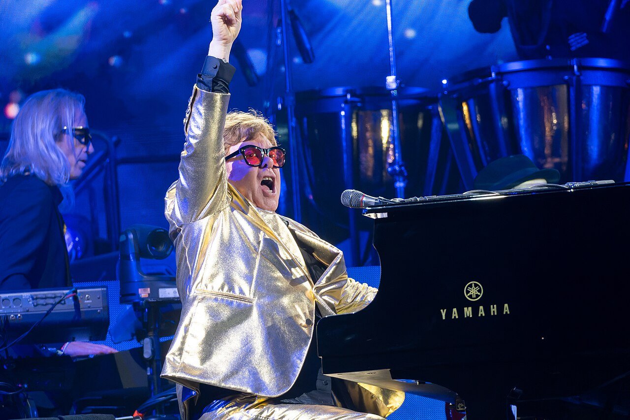 Elton John in a gold jacket raising one arm while performing at a piano on stage at Glastonbury in 2023