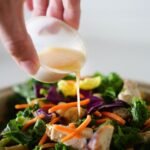 Hand pouring healthy salad dressing from a small cup over a bowl of greens and carrots