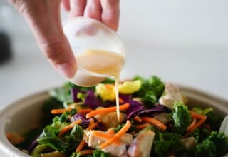 Hand pouring healthy salad dressing from a small cup over a bowl of greens and carrots