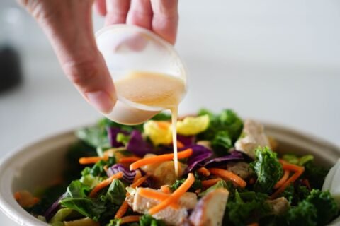 Hand pouring healthy salad dressing from a small cup over a bowl of greens and carrots