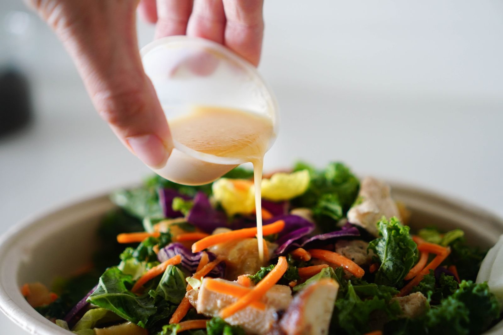 Hand pouring healthy salad dressing from a small cup over a bowl of greens and carrots