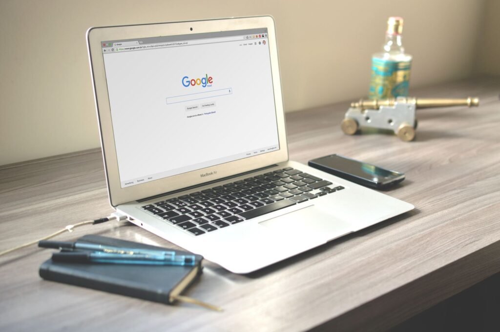 Laptop open to the Google search page on a wooden desk next to a smartphone and notebook