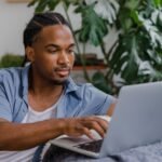 Young man in a blue shirt working on a laptop at a home desk beside a large monstera plant