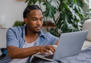 Young man in a blue shirt working on a laptop at a home desk beside a large monstera plant