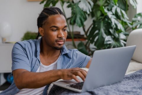 Young man in a blue shirt working on a laptop at a home desk beside a large monstera plant